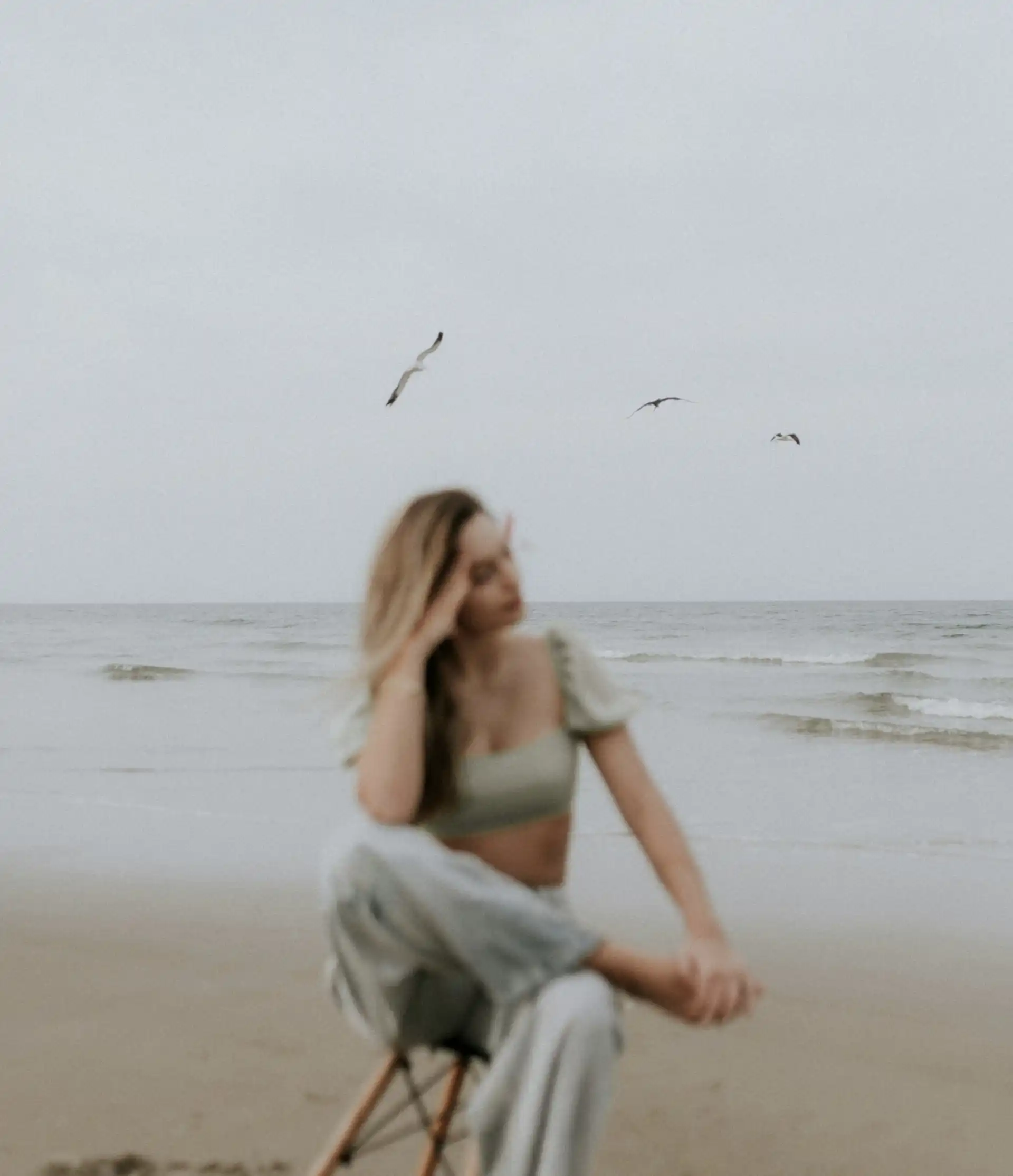 Photo floue représentant une femme assise sur une chaise, sur la plage, jambes croisées, qui semble pensive, avec trois oiseaux en arrière plan.