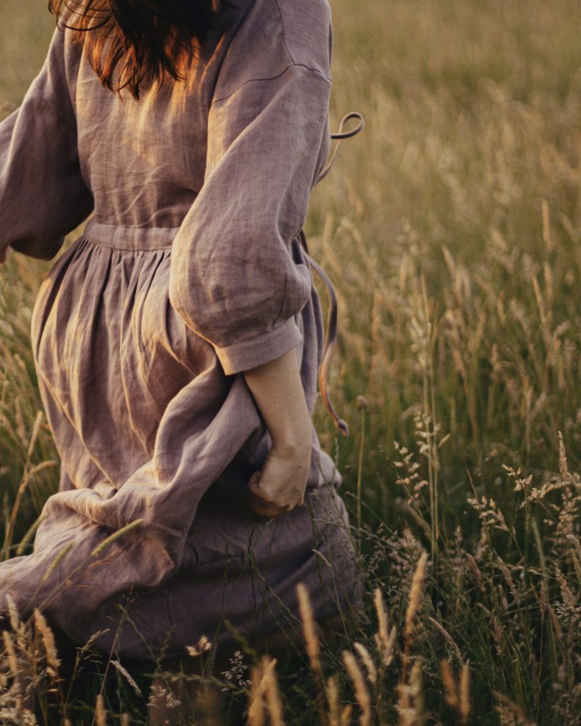 Photo représentant une femme en robe (en lin) en train de courir librement dans un pré de hautes herbes, le tout baigné d'une lumière dorée.