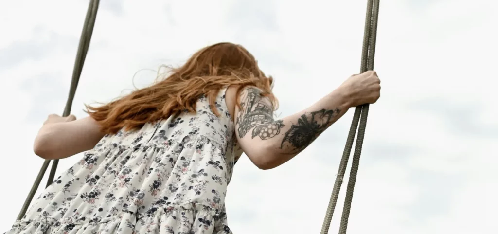 Photo d'une femme portant des tatouages sur le bras, une robe blanche à petites fleurs bleues, en train de faire de la balançoire.