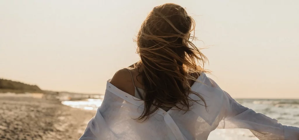 Photo représentant le haut du corps d'une femme de dos, cheveux au vent, en train de marcher bras ouverts sur la plage, avec une lumière dorée.