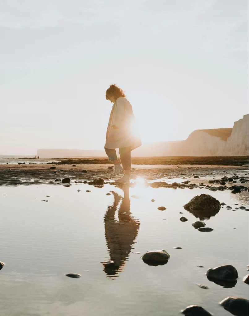 Photo représentant une femme en train de marcher sur la plage, avec des falaises et un début de coucher de soleil en arrière plan.