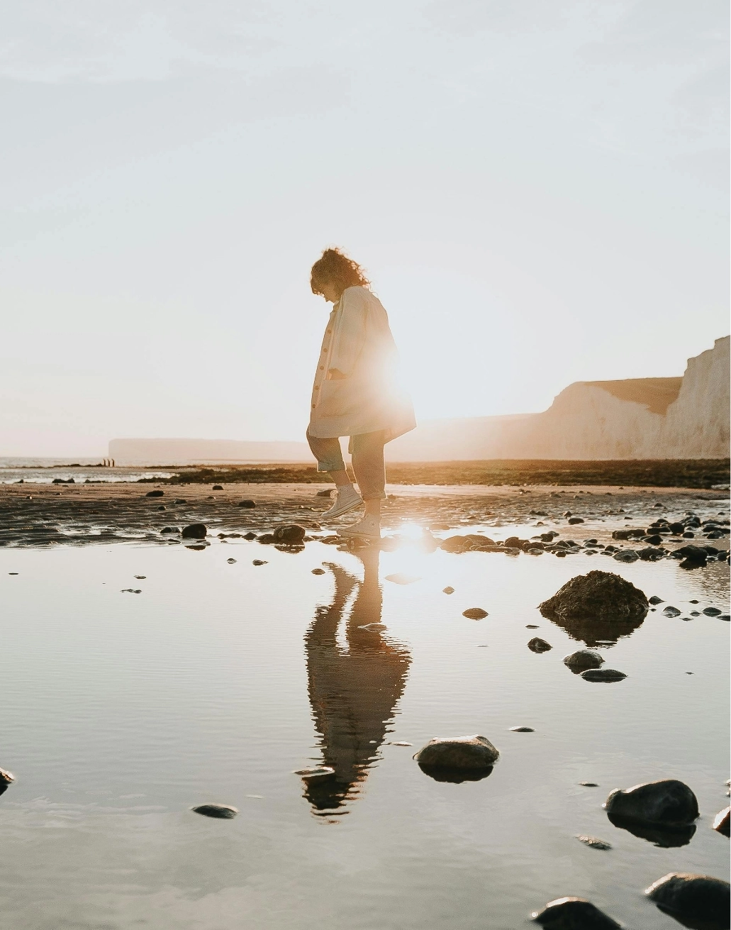 Photo représentant une femme en train de marcher sur la plage, avec des falaises et un début de coucher de soleil en arrière plan.